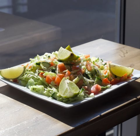 Fresh green salad with tomatoes, peppers, and lime wedges on a white square plate by a window
