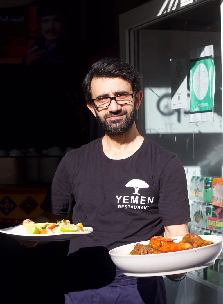 Man wearing Yemen Restaurant t-shirt holding two plates of food with vegetables and meat dishes